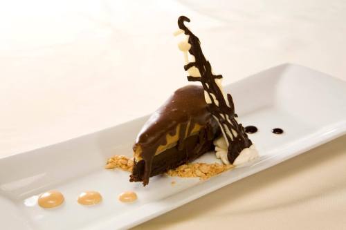 A slice of chocolate dessert with a creamy filling, garnished with chocolate curls and drizzles on a white plate.