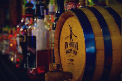 A wooden barrel with "Idyll Mountain Distillery" engraved, surrounded by various bottles of liquor on a bar.