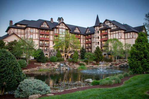 A picturesque hotel with a European-style facade, surrounded by lush greenery and a tranquil pond with a fountain.