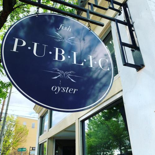Sign for "P.U.B.L.I.C" featuring "fish" and "oyster" on a blue background, hanging outside a restaurant.