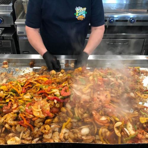 A chef in gloves stirs a large skillet of colorful sautéed vegetables and shrimp in a busy kitchen. Steam rises.
