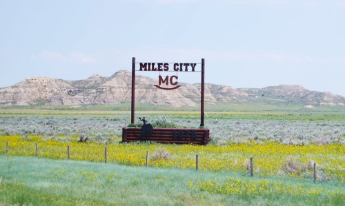 Sign for Miles City, Montana, with rolling hills and wildflowers in the foreground under a clear blue sky.