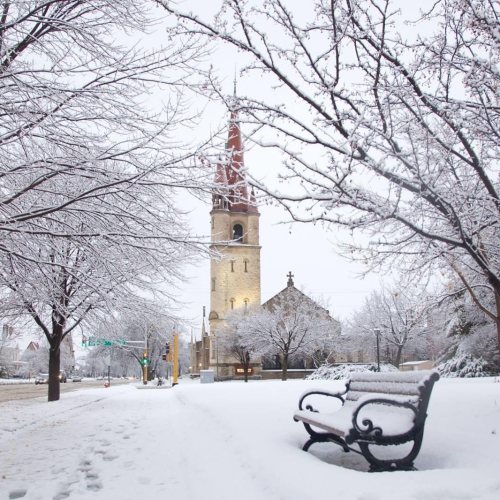 A snowy scene featuring a church tower, bare trees, and a bench in a winter landscape.