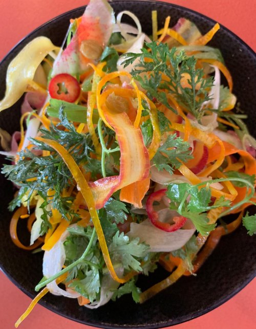 A vibrant salad with colorful vegetable ribbons, herbs, and sliced chili in a black bowl on a bright orange background.