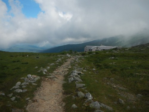 A rocky path leads through green grass towards a building under a cloudy sky in a mountainous landscape.