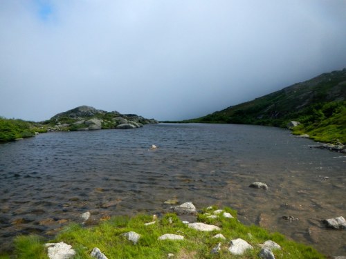 A serene lake surrounded by rocky hills and lush greenery under a cloudy sky.