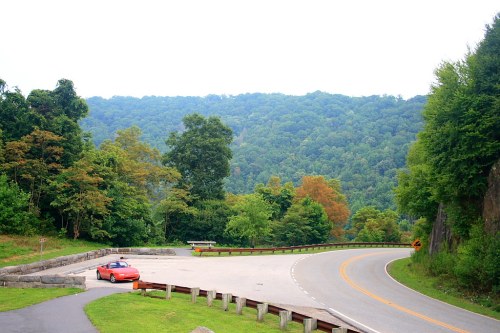 A red convertible parked at a scenic overlook with lush green hills in the background and a winding road.