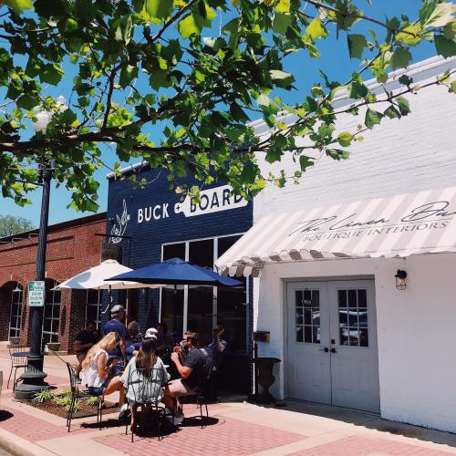 A sunny street scene featuring outdoor seating at a café, with trees and buildings in the background.