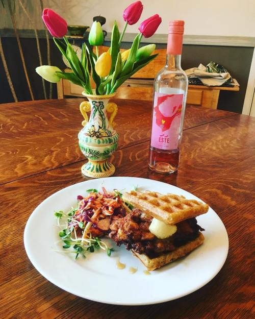 A plate with a waffle sandwich filled with fried chicken, a side salad, a vase of tulips, and a bottle of rosé wine.