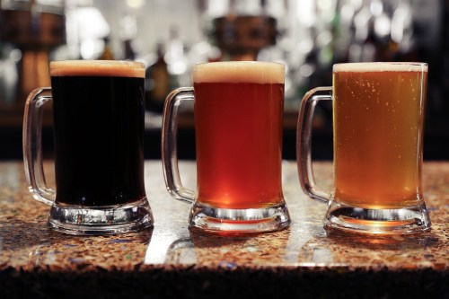Three mugs of beer on a bar counter: a dark stout, a reddish amber ale, and a light golden lager.