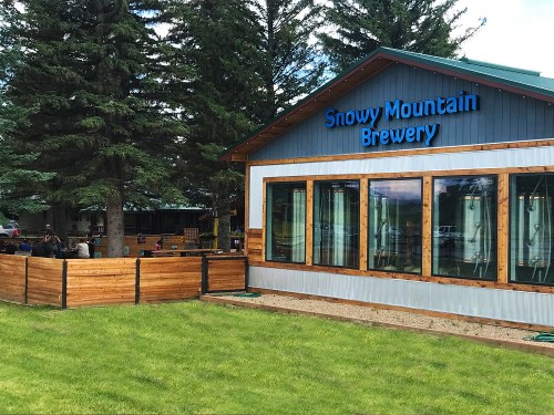 Exterior of Snowy Mountain Brewery, featuring large windows, wooden accents, and a grassy area with trees.