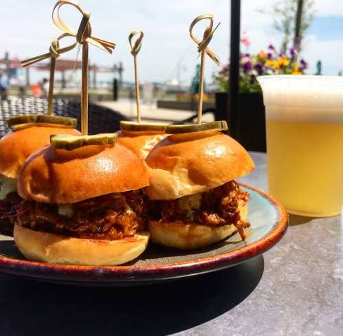 Three pulled pork sliders with pickles on a plate, accompanied by a glass of beer, set against a sunny outdoor backdrop.