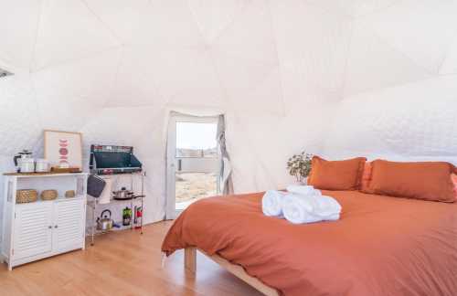 Cozy interior of a dome-shaped room with a large bed, white towels, and a small kitchenette area. Natural light enters through the door.