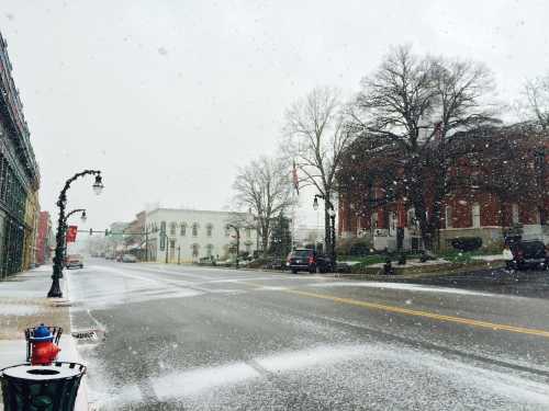 A snowy street scene with buildings, trees, and parked cars, creating a quiet, wintry atmosphere.