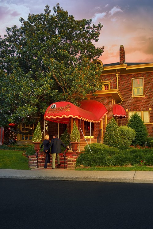 A couple stands under a red awning in front of a brick house, surrounded by greenery at sunset.