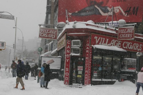A snowy street scene featuring a storefront labeled "Village Cigars" with people walking in heavy snowfall.