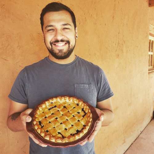 A smiling man holds a freshly baked pie with a lattice crust, standing against a rustic wall.