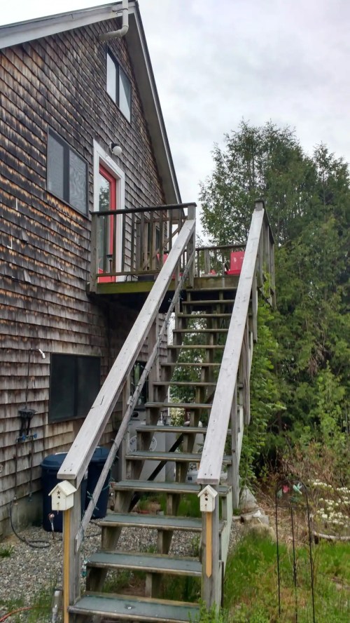 A wooden staircase leads up to a house with a shingled exterior and a red door, surrounded by greenery.
