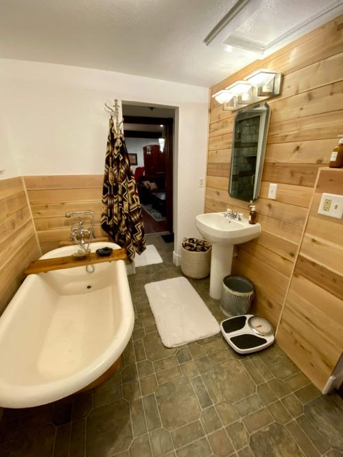 Cozy bathroom featuring a vintage tub, wooden walls, sink, and decorative elements. Soft lighting enhances the ambiance.