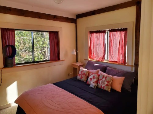 Cozy bedroom with a black bedspread, colorful pillows, and large windows with red curtains letting in natural light.
