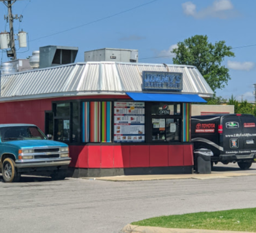 A colorful drive-thru restaurant with a silver roof, featuring a blue truck and a black vehicle parked outside.