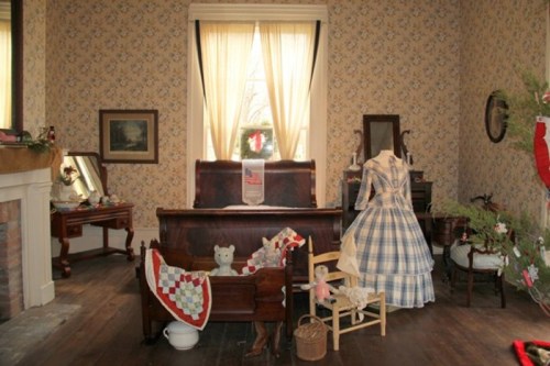 A vintage bedroom with a wooden bed, quilt, dress form, and decorative items, featuring floral wallpaper and natural light.