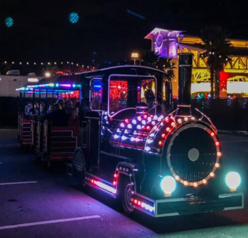 A brightly lit miniature train with colorful lights, parked at night with festive decorations in the background.