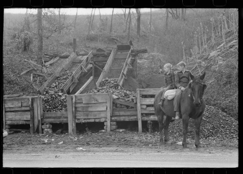 Two children sit on a horse near a coal loading area with piles of coal and wooden structures in a rural setting.