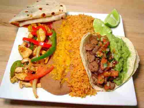 A plate of Mexican food featuring tacos, rice, beans, grilled vegetables, and lime wedges.