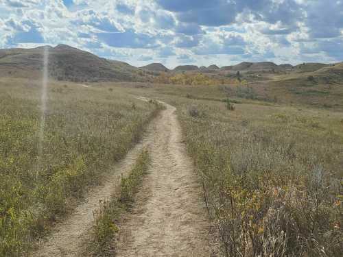 A winding dirt path through grassy hills under a cloudy sky, with hints of autumn foliage in the distance.