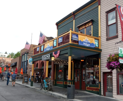A colorful street scene featuring a bar with flags, outdoor seating, and people walking by in a lively atmosphere.