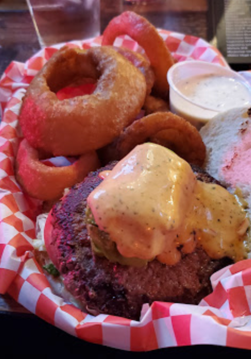 A juicy burger topped with sauce, served with onion rings and a side of dipping sauce in a red-checkered basket.