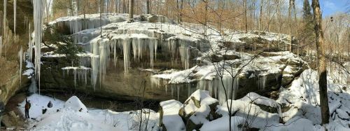 A snowy landscape featuring a rocky cliff with icicles hanging down, surrounded by bare trees.