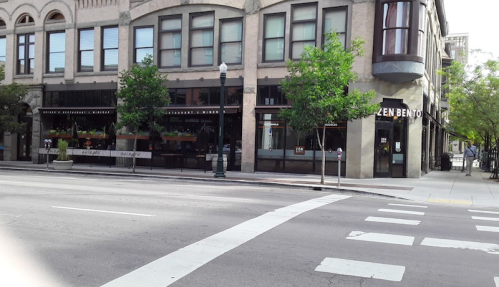 A street view of a restaurant named "Zen Bento" with outdoor seating and greenery along the sidewalk.