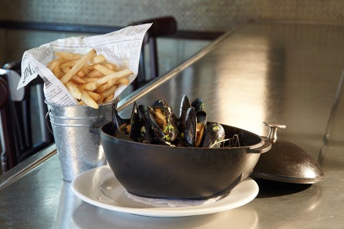 A black pot filled with mussels sits on a plate next to a metal cup of French fries wrapped in paper.