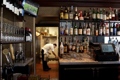 A bar with shelves of liquor bottles, a bartender organizing glasses, and a kitchen visible in the background.
