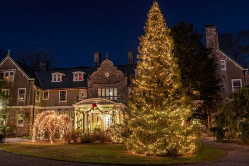 A beautifully lit Christmas tree stands in front of a decorated mansion at night, surrounded by festive lights.