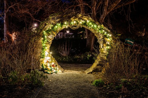 A stone archway adorned with glowing lights, surrounded by bare trees and a dark garden path at night.