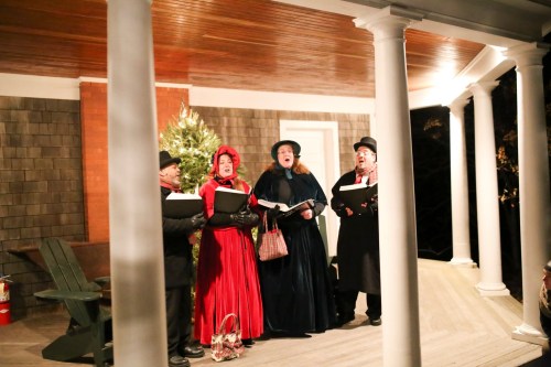 A group of four carolers in festive attire singing on a porch decorated for the holidays.