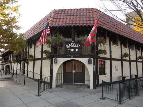 Exterior of the Basque Center, featuring a tiled roof, flags, and decorative plants.