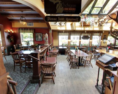 Cozy restaurant interior with wooden tables and chairs, large windows, and a "Please Seat Yourself" sign.
