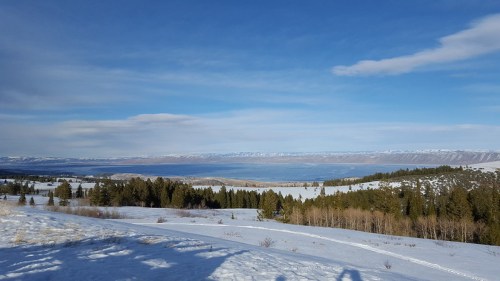 A panoramic view of a snowy landscape with trees, mountains, and a lake under a clear blue sky.