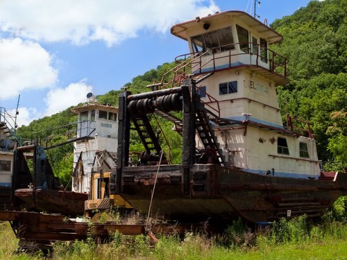 Abandoned tugboats on land, surrounded by overgrown grass and trees under a blue sky.