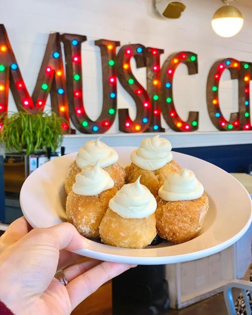 A hand holds a plate of six cream-topped pastries in front of a colorful "MUSIC CITY" sign.