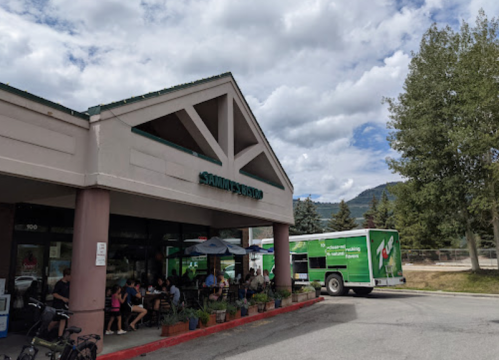 A café with outdoor seating, people dining, and a green delivery truck parked nearby under a cloudy sky.