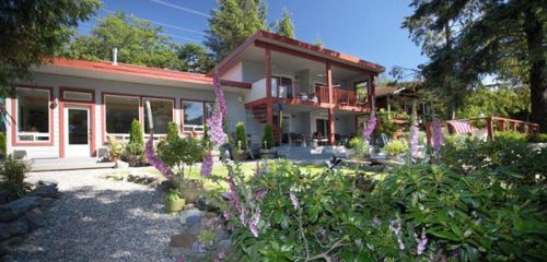 A modern two-story house surrounded by greenery and flowers, with a clear blue sky in the background.