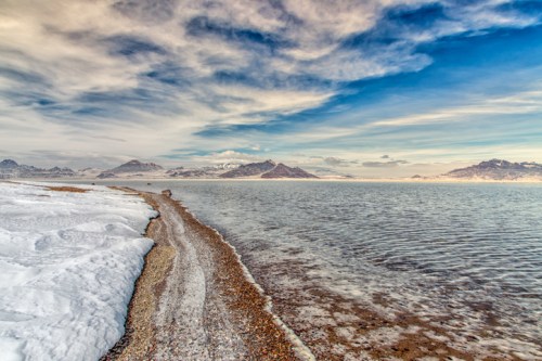 A serene landscape featuring a snowy shoreline meeting a calm body of water, with mountains in the background under a blue sky.