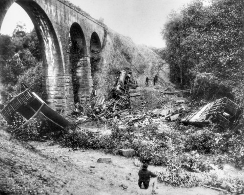 A historic black-and-white photo of a train wreck near a stone bridge, with debris scattered and people inspecting the scene.