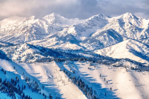 Snow-covered mountains with peaks and valleys, casting long shadows under a cloudy sky.
