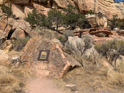 A rocky landscape with a plaque on a boulder, surrounded by sparse vegetation and towering cliffs in the background.
